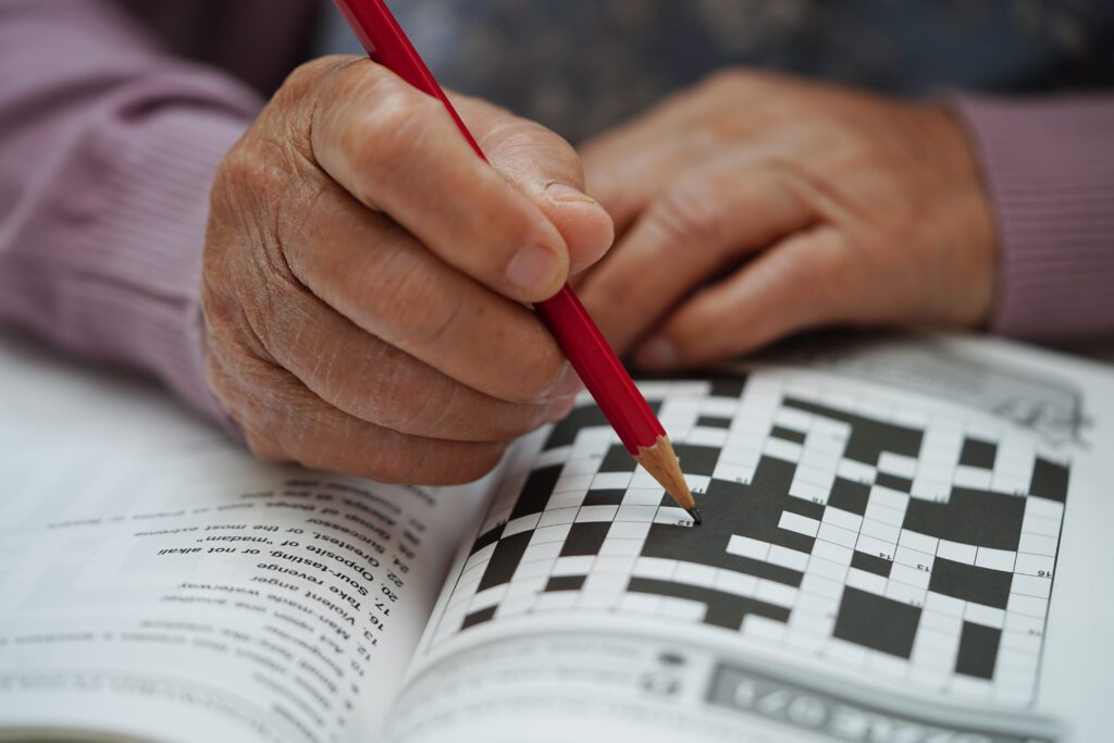 A person holding a red pencil is filling in answers on a printed crossword puzzle in a magazine or book—a classic example of brain games for seniors. The focus is on their hands and the puzzle.