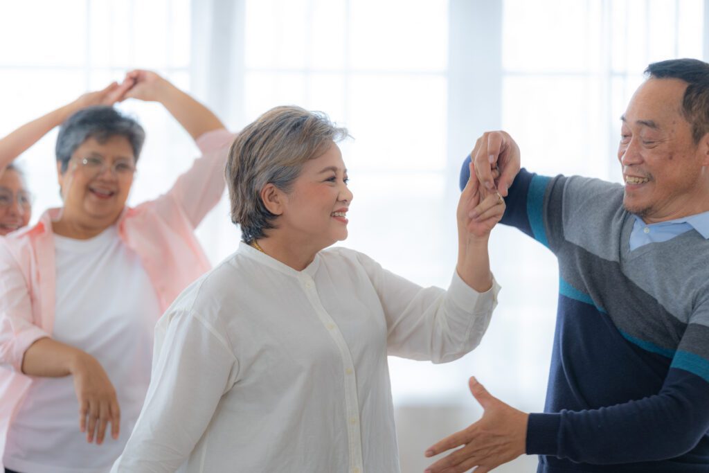 Two older adults smile and dance together indoors, enjoying the benefits of dancing, while another pair moves in the background. The room is bright with large windows, creating a cheerful atmosphere.