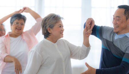 Two older adults smile and dance together indoors, enjoying the benefits of dancing, while another pair moves in the background. The room is bright with large windows, creating a cheerful atmosphere.