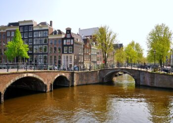 Amsterdam reveals its charm along a scenic canal, where a brick bridge connects historic narrow houses and trees line the water under a clear sky. People are walking and cycling across the bridge, soaking in the vibrant atmosphere.