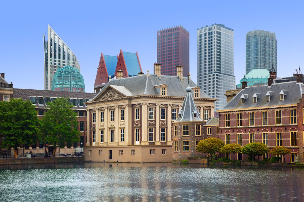 Amsterdam reveals a striking contrast as the historic buildings of the Binnenhof and Mauritshuis are reflected in a pond, with modern skyscrapers rising in the background under The Hague's clear blue sky.