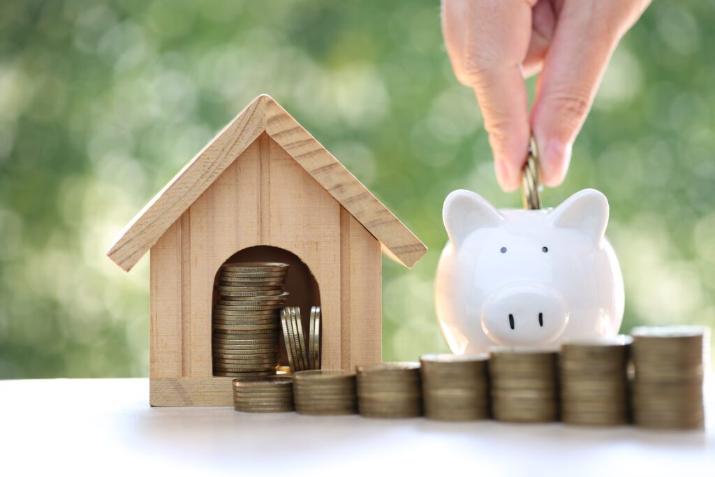 A hand places a coin into a white piggy bank next to a wooden house model filled with coins, symbolizing savings and common mortgage mistakes homeowners make. Stacks of coins are lined up on a white surface with a blurred green background.