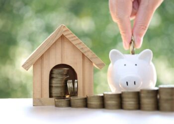 A hand places a coin into a white piggy bank next to a wooden house model filled with coins, symbolizing savings and common mortgage mistakes homeowners make. Stacks of coins are lined up on a white surface with a blurred green background.