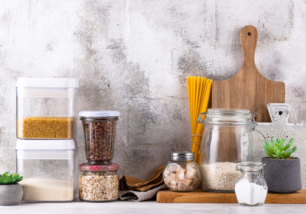 Various kitchen storage containers, jars with pasta, rice, oats, and sugar cubes, a wooden cutting board, a small succulent plant, and salt on a countertop against a light textured wall.