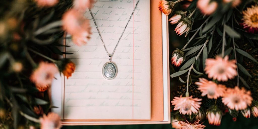 A silver oval pendant necklace, one of a kind jewelry designed by you, rests on a handwritten letter inside an open box, surrounded by pink and peach flowers with green leaves.