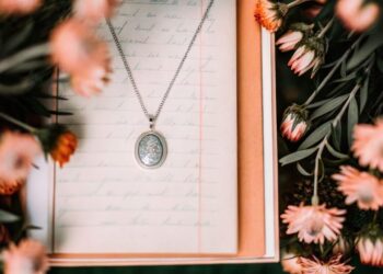 A silver oval pendant necklace, one of a kind jewelry designed by you, rests on a handwritten letter inside an open box, surrounded by pink and peach flowers with green leaves.