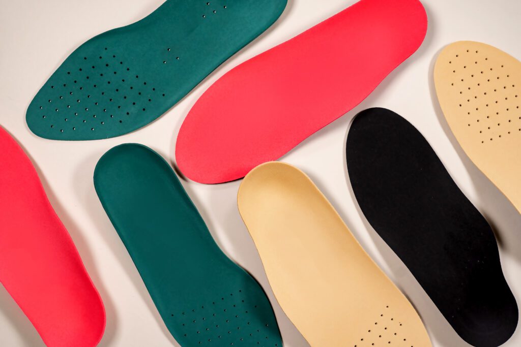 Several colorful shoe insoles, ideal as orthotics for seniors, are arranged on a light background. Available in green, red, beige, and black, some feature small perforated holes and vary slightly in shape and size.
