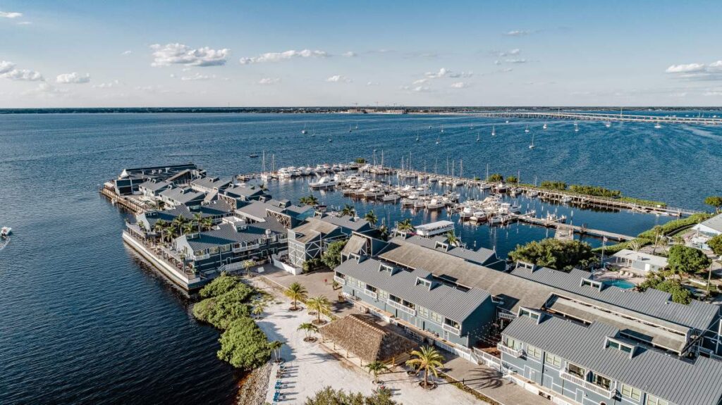 Aerial view of a marina with numerous boats docked, surrounded by buildings with metal roofs. The marina is situated on a large body of water under a blue sky with scattered clouds.