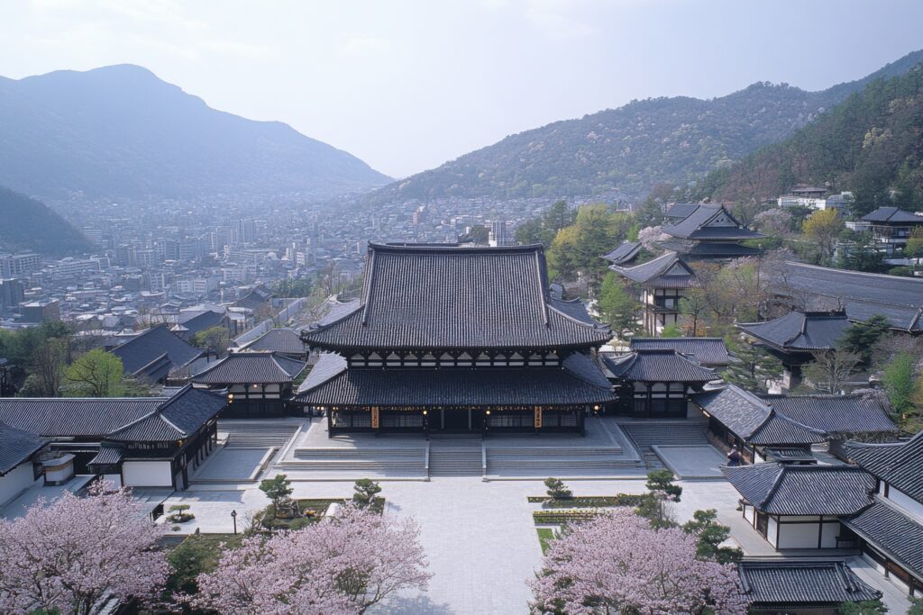 Traditional Japanese temple complex surrounded by blooming cherry blossom trees, with mountains and a cityscape visible in the background under a clear sky.
