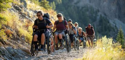 A group of people using wheelchairs enjoy hiking together on a rocky trail through a scenic, sunlit mountain landscape, showcasing accessible group travel for seniors and travelers with disabilities amidst trees and lush greenery.