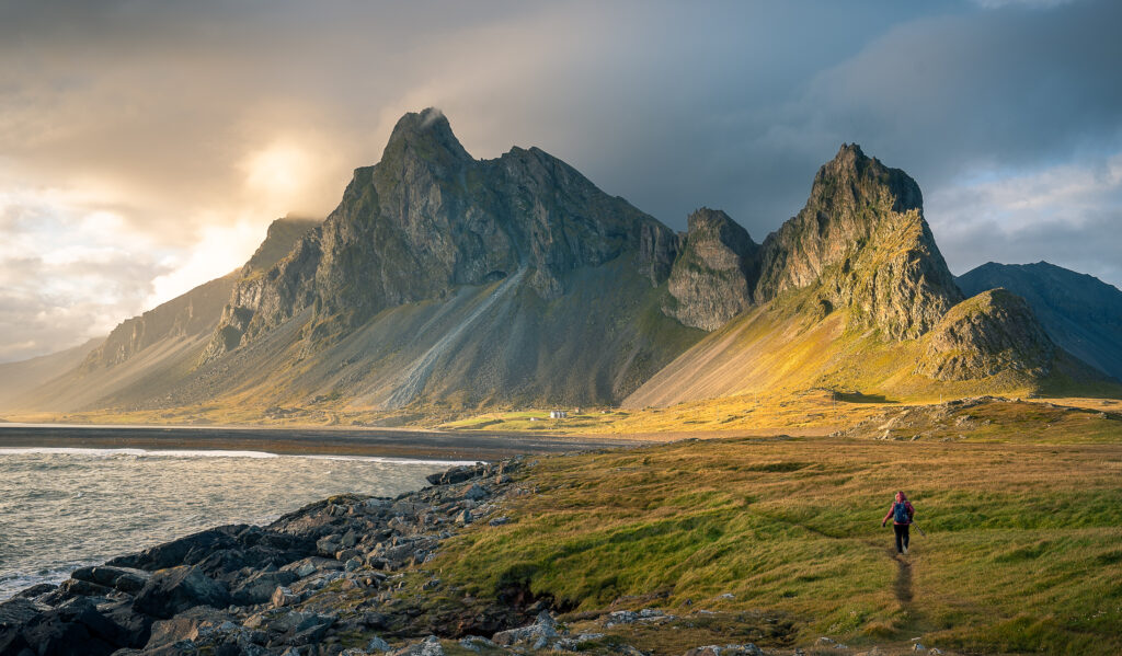 A person with a backpack walks along a grassy coastline toward dramatic, jagged mountains under a cloudy, golden-lit sky in a remote, scenic landscape.