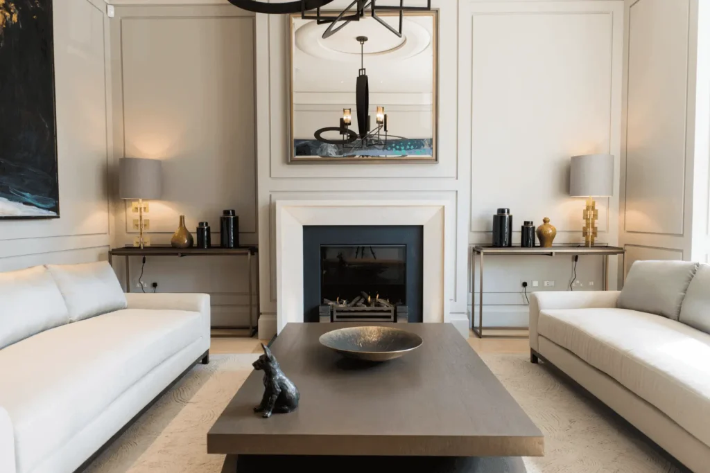A modern living room with two white sofas, a dark wood coffee table holding a decorative bowl and black dog figurine, a fireplace, large mirror, two table lamps, and neutral walls with subtle paneling—perfect for staging tips inspiration.