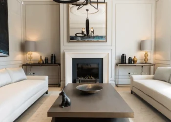 A modern living room with two white sofas, a dark wood coffee table holding a decorative bowl and black dog figurine, a fireplace, large mirror, two table lamps, and neutral walls with subtle paneling—perfect for staging tips inspiration.