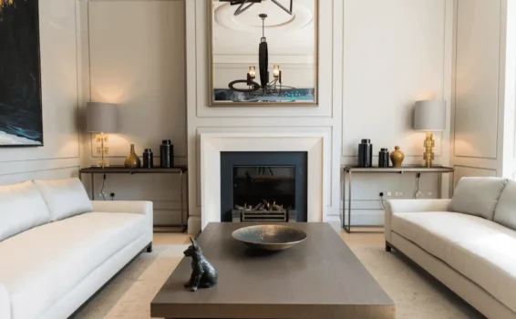 A modern living room with two white sofas, a dark wood coffee table holding a decorative bowl and black dog figurine, a fireplace, large mirror, two table lamps, and neutral walls with subtle paneling—perfect for staging tips inspiration.