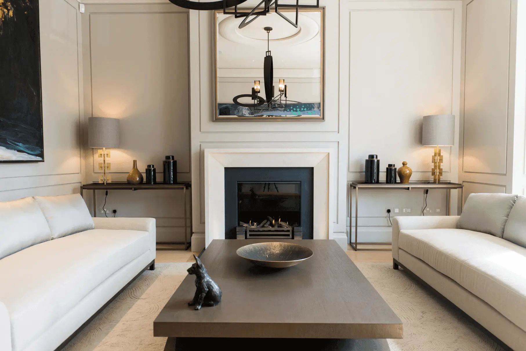 A modern living room with two white sofas, a dark wood coffee table holding a decorative bowl and black dog figurine, a fireplace, large mirror, two table lamps, and neutral walls with subtle paneling—perfect for staging tips inspiration.