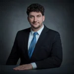A man with curly brown hair and a beard, wearing a black suit, light blue dress shirt, and blue tie, sits at a table with a neutral gray background, smiling slightly at the camera.