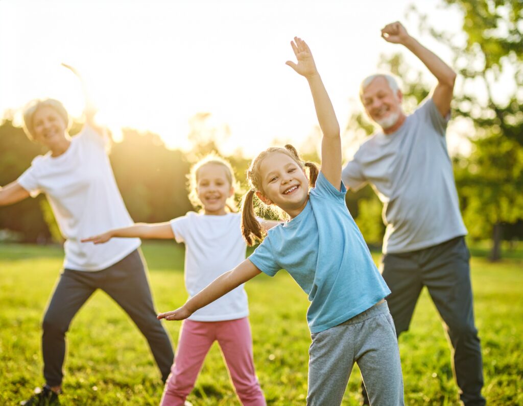 Two children and two adults smile and stretch their arms while exercising together outside on a sunny day—it's FITNESS FUN WITH GRANDKIDS, enjoying movement on green grass with trees in the background.