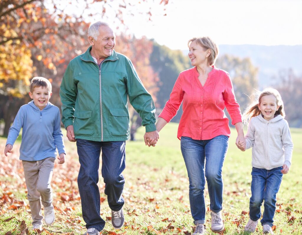Two adults and two children walk hand-in-hand outdoors on a sunny day, smiling and surrounded by autumn leaves—a moment that beautifully captures the health benefits of being a grandparent amid nature’s cheerful embrace.