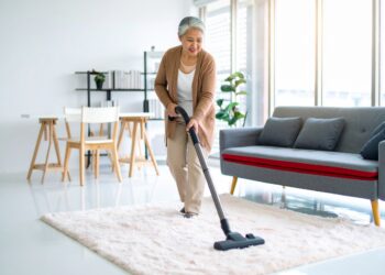 An older woman smiles while vacuuming a white rug in a bright, modern living room—she’s enjoying one of the best vacuums, featured among the Favorite Vacuums of 2025.