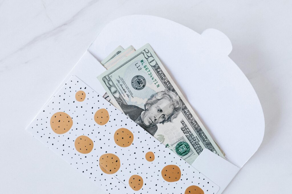 A white envelope with gold polka dots, perfect for gifting money to family, contains several U.S. twenty-dollar bills, partially visible against a white background.