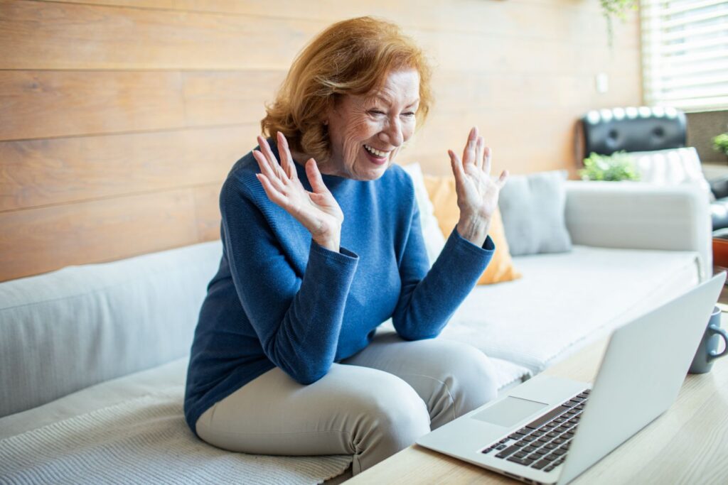 An older woman with red hair, wearing a blue sweater and beige pants, sits on a couch smiling and waving at a laptop during a video call, sharing Long-Distance Grandparenting Tips to Stay Close from her cozy, sunlit living room.
