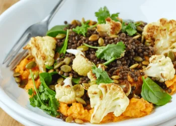 A white bowl contains anti-inflammatory foods for seniors like roasted cauliflower, black lentils, spinach, cilantro, pumpkin seeds, and mashed sweet potatoes, with a fork resting at the side.