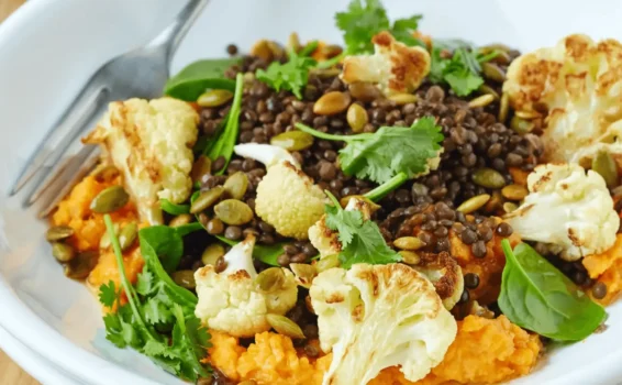 A white bowl contains anti-inflammatory foods for seniors like roasted cauliflower, black lentils, spinach, cilantro, pumpkin seeds, and mashed sweet potatoes, with a fork resting at the side.