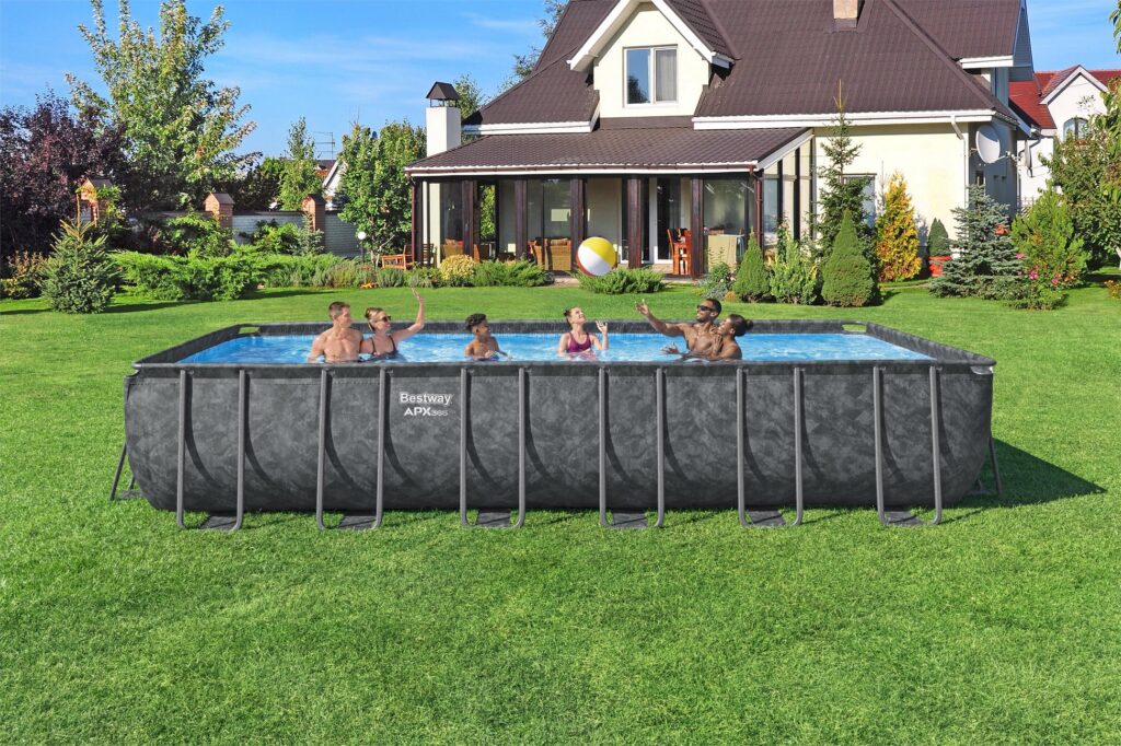 Five people play with a beach ball in the best backyard pool—a large rectangular above-ground pool set up in a grassy backyard with a house and trees in the background on a sunny day.