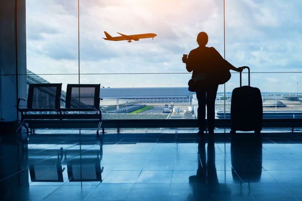 A silhouette of a person holding a coffee cup and standing with luggage by large windows at an airport, watching an airplane take off against a cloudy sky.