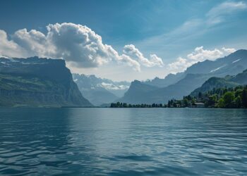 A tranquil lake is surrounded by lush green hills and snow-capped mountains under a partly cloudy sky. Trees line the shoreline, and mountain peaks rise dramatically in the background.