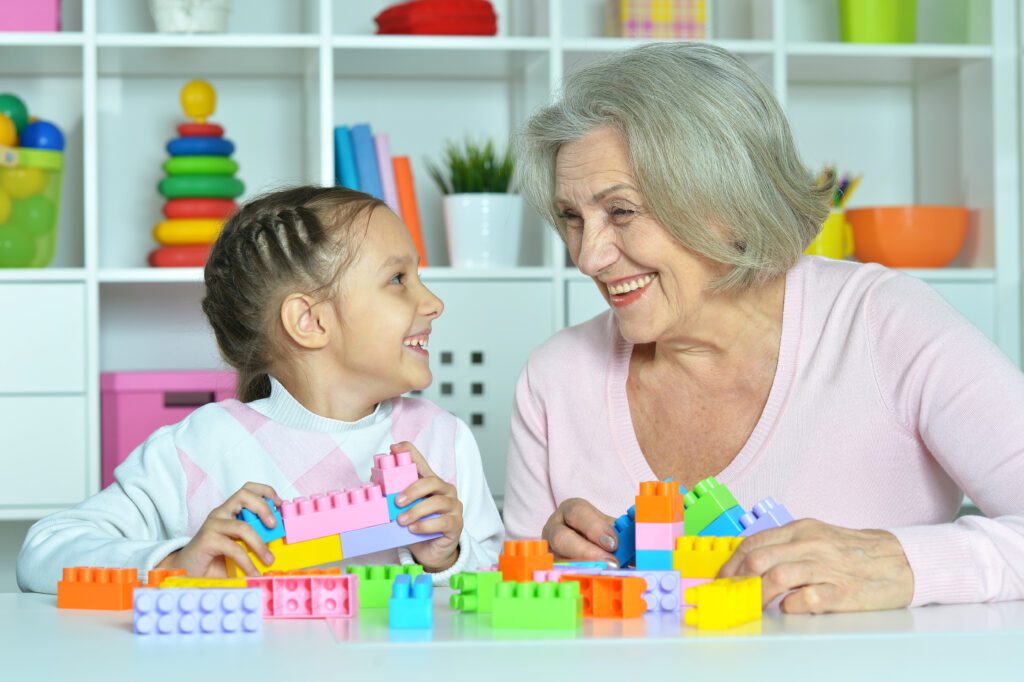A smiling elderly woman and a young girl sit at a table building LEGO with Grandkids sets, sharing a joyful moment. Shelves with toys and decorations are visible in the background.