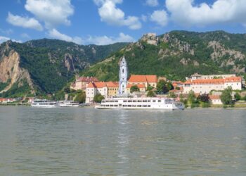 A scenic riverside town with a tall church tower, colorful buildings, and green hills in the background. Two white river cruise boats are docked along the water under a partly cloudy sky.