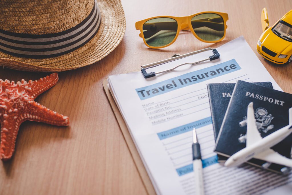 A travel insurance form on a clipboard, with passports, a toy airplane, a pen, a straw hat, sunglasses, a toy car, and a starfish arranged on a wooden table—ideal for those seeking travel insurance for seniors.