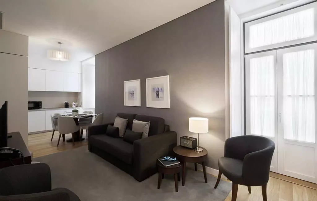 Modern living room with a dark sofa, armchair, side tables, and lamps. There are two framed pictures on a gray wall, a dining table with chairs, and a small kitchen area with white cabinets in the background. Large windows let in light.