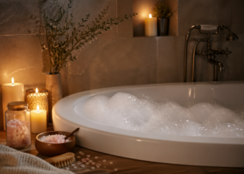 A bubble-filled bathtub surrounded by lit candles, a jar and bowl of pink bath salts, a brush, and green branches, creating a relaxing spa-like atmosphere in a softly-lit bathroom.