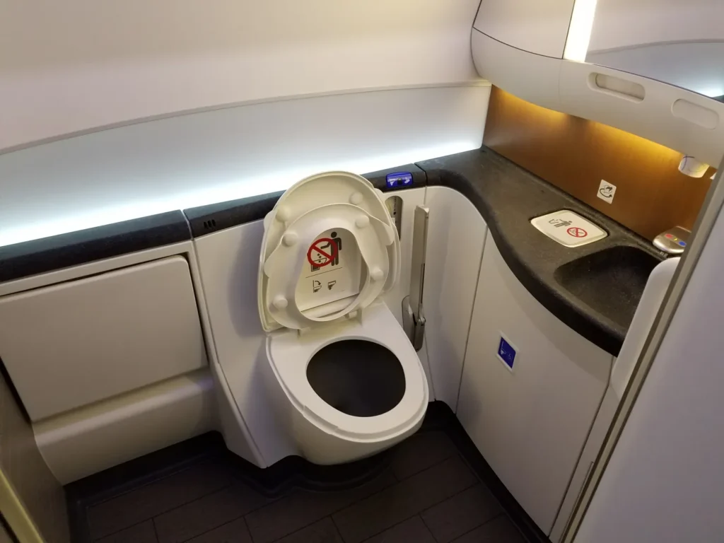 An airplane lavatory with a toilet, open lid showing a no-smoking sign, a sink with a no-smoking symbol on the counter, modern lighting and surfaces—designed for convenience and accessibility, offering helpful airplane bathroom tips for wheelchair users.