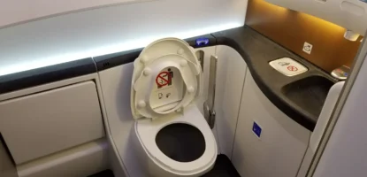 An airplane lavatory with a toilet, open lid showing a no-smoking sign, a sink with a no-smoking symbol on the counter, modern lighting and surfaces—designed for convenience and accessibility, offering helpful airplane bathroom tips for wheelchair users.