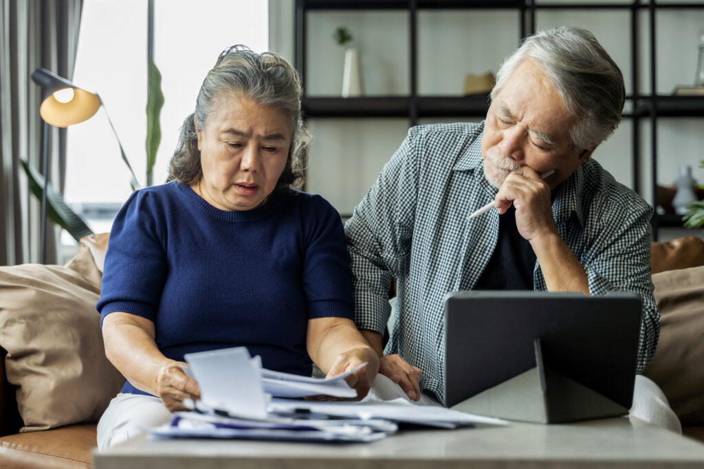 An older couple sits on a couch, looking concerned while reviewing documents about reverse mortgage myths. The woman holds papers, and the man holds a pen, glancing at a tablet on the table in front of them.