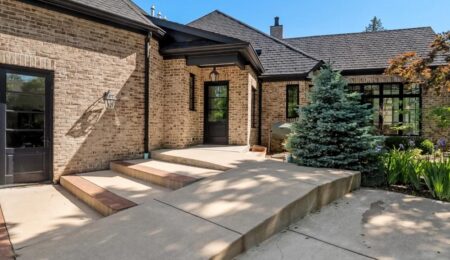 A modern brick house with large black-framed windows, a glass door, and a wide concrete entryway with steps. Landscaping includes a small evergreen tree and green plants under a clear blue sky.