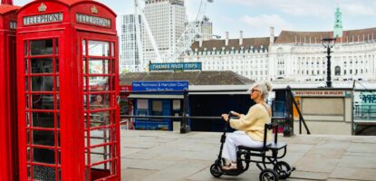 An elderly woman rides a mobility scooter past two classic red British telephone boxes near the Thames River in London, one of the best accessible cities in Europe, with the London Eye and city buildings in the background.
