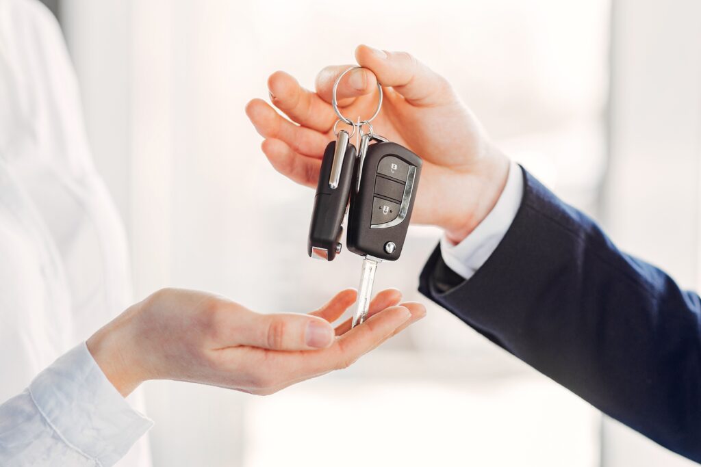 A person in a suit hands over car keys to another, symbolizing the process of how to transfer a car title after death. The background is bright and softly blurred.