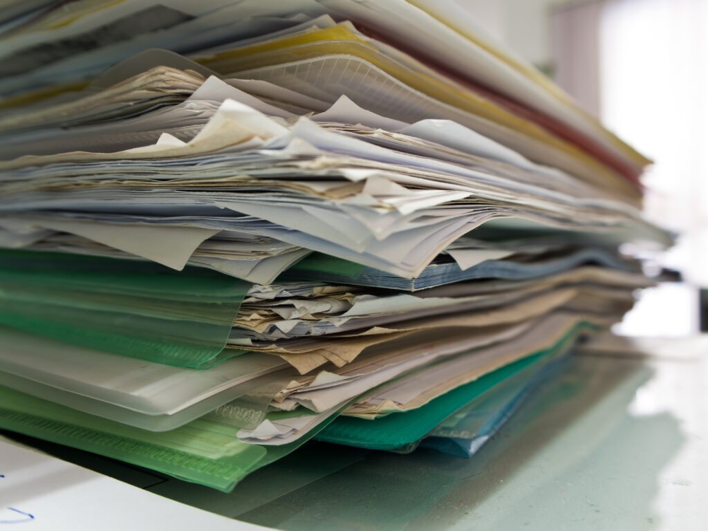 A large, messy stack of assorted papers and folders sits on a desk, suggesting disorganization or a heavy workload in an office environment.