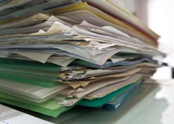 A large, messy stack of assorted papers and folders sits on a desk, suggesting disorganization or a heavy workload in an office environment.