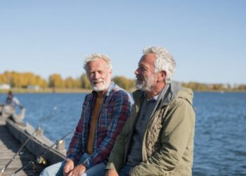 Two older men sit on a wooden dock by a lake with fishing rods, talking and smiling. Surrounded by autumn foliage and a blue sky, they enjoy a perfect day of fishing for beginners and pros alike.