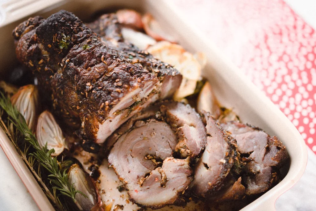 A roasted rolled porketta shoulder is sliced and arranged in a baking dish with fresh rosemary and roasted onions, seasoned with savory herbs and spices.