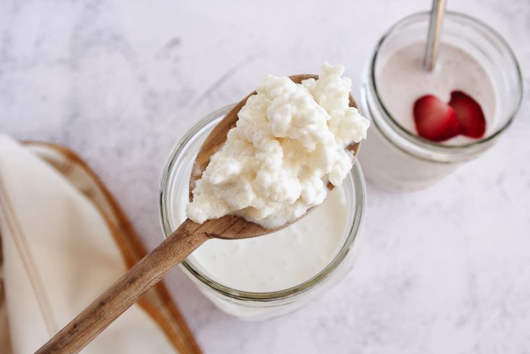 A wooden spoon holding creamy cottage cheese over a glass jar, with another jar containing yogurt and strawberry slices in the background—perfect for enjoying kefir benefits alongside wholesome dairy treats.