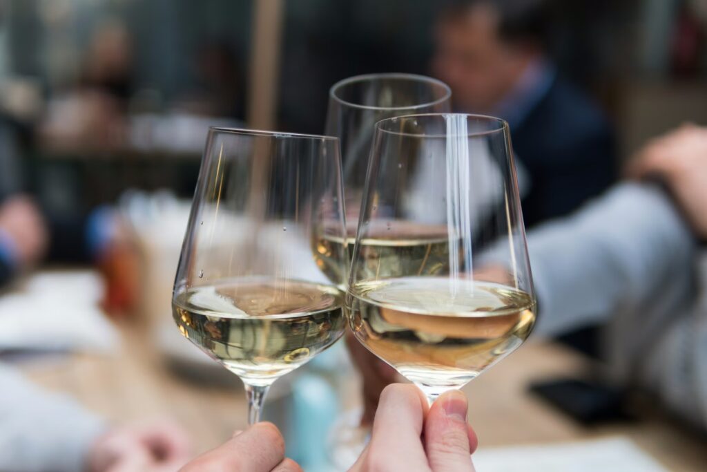 Three people clink glasses of low-alcohol wine in a toast at a table, with blurred figures and a softly lit background suggesting a relaxed social gathering.