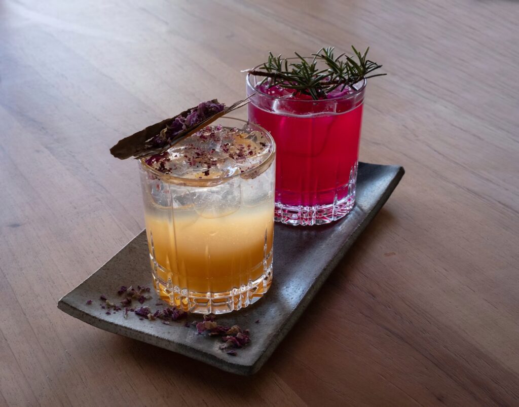 Two colorful drinks made with low-alcohol wine in clear glasses sit on a rectangular ceramic tray. One drink is orange with ice and a bay leaf, while the other is bright pink with ice and topped with rosemary.