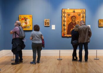 Visitors view religious icon paintings displayed on blue walls in an art gallery. Four people stand observing the artwork, which includes several small and one large, colorful religious icons. The gallery has wooden floors.