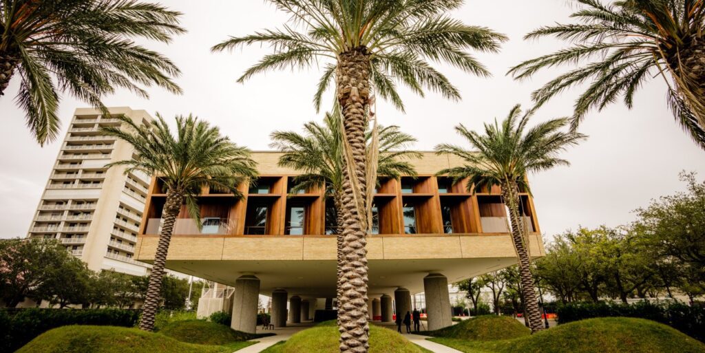 Modern building with large windows and a raised structure supported by columns, surrounded by palm trees and green grass beneath a cloudy sky. The International African American Museum Charleston stands out, with a tall apartment building in the background.